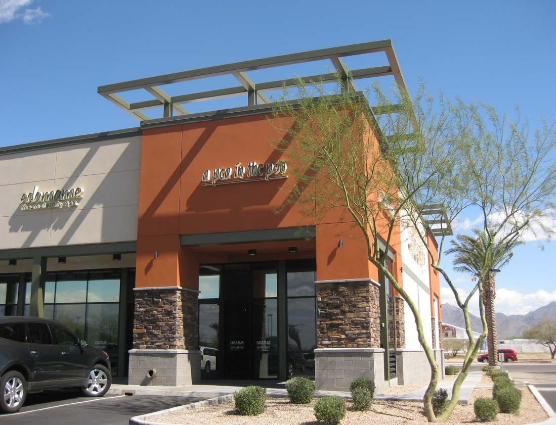 Corner retail entry at Chauncey Ranch with a steel trellis canopy, stone base, and orange stucco volume against the Arizona sky