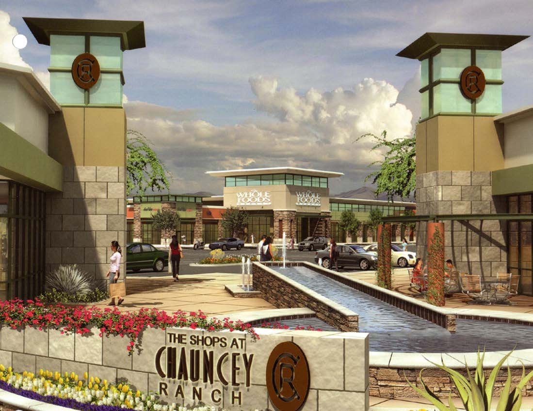 Entry court at The Shops at Chauncey Ranch with the stone monument sign, water feature, palms, and flanking retail towers