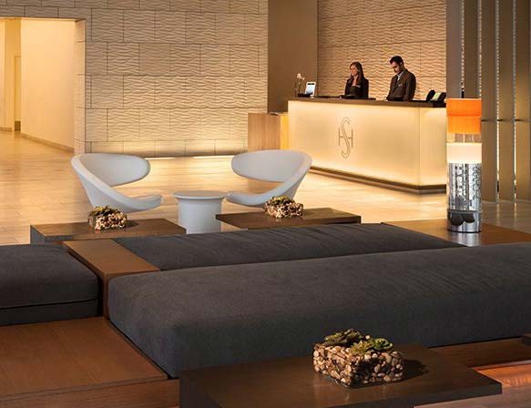 Shore Hotel reception desk in backlit stone with two staff members, white lounge chairs, and a low upholstered bench in the foreground