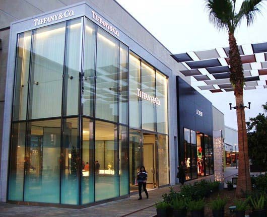 Two-story glazed corner storefront at University Town Center with a slatted canopy and palm trees along the open-air walkway