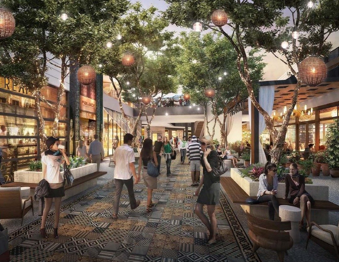 Open-air retail promenade at Westfield Century City with mature trees, pendant lighting, patterned paving, and shoppers seated in the landscaped central plaza